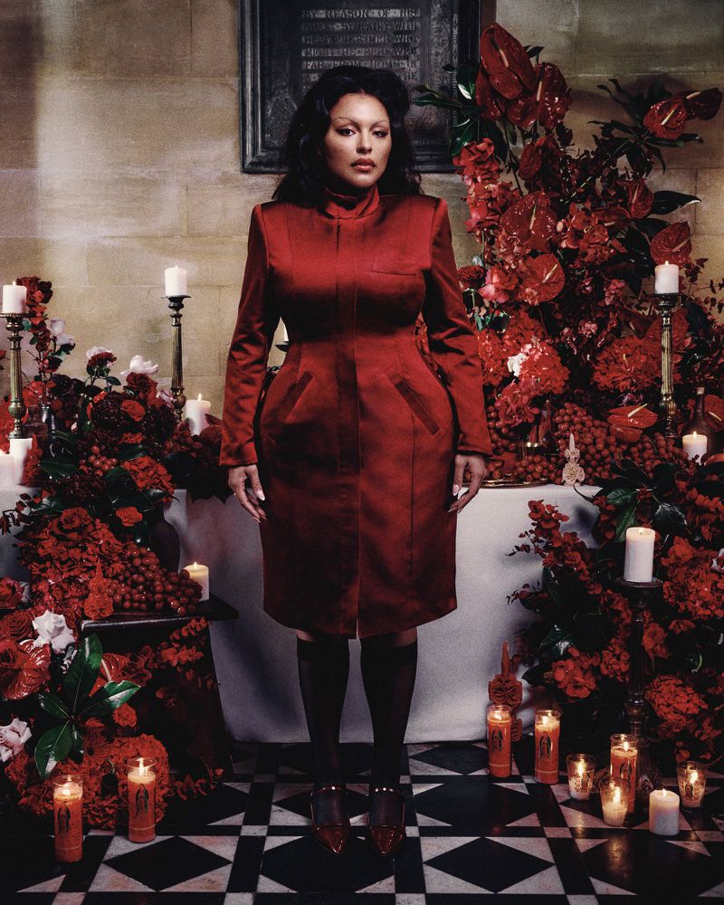 Elegant woman in a red dress stands surrounded by lush red flower arrangements and lit candles, captured in a full-length shot.