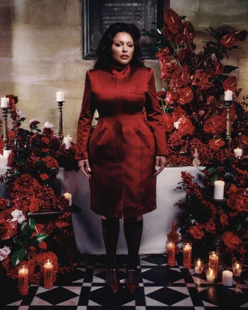 Elegant woman in a red dress stands surrounded by lush red flower arrangements and lit candles, captured in a full-length shot.