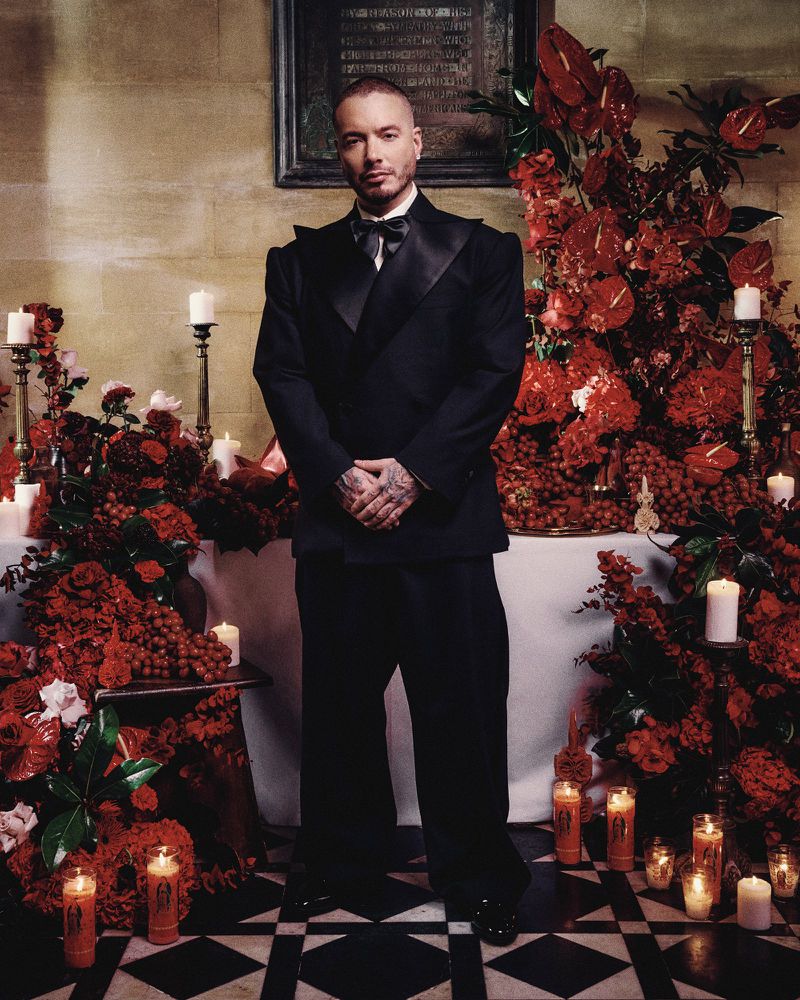 Full-length portrait of adult male, J Balvin, in a black tuxedo surrounded by red floral arrangements and candles. Formal, indoor setting.