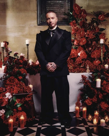 Full-length portrait of adult male, J Balvin, in a black tuxedo surrounded by red floral arrangements and candles. Formal, indoor setting.