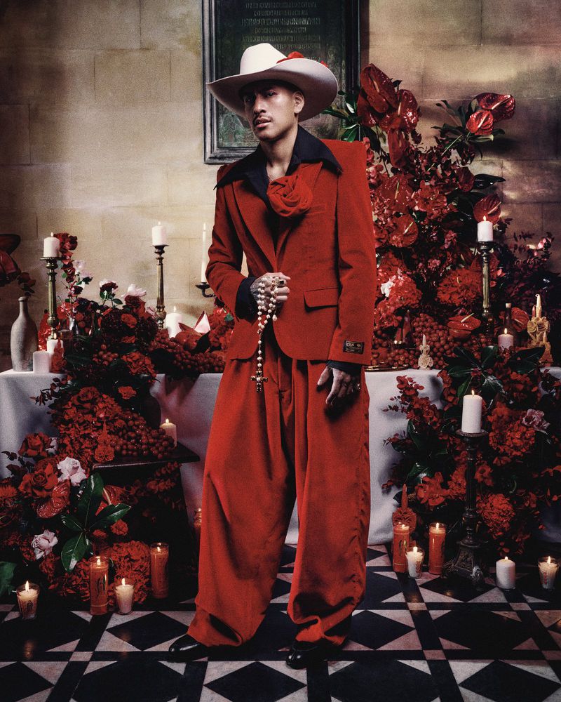 A person in a red suit and white cowboy hat stands among red flowers and candles in an indoor setting with black and white tiled floor.