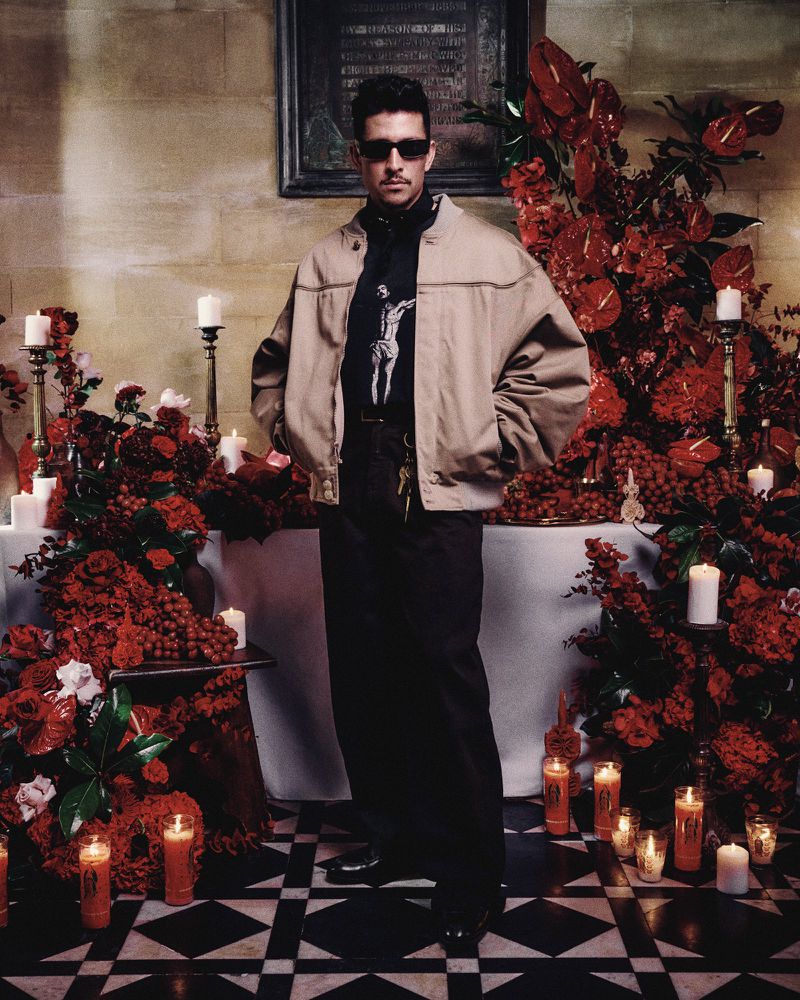 Full shot of a man in a jacket and sunglasses, posing in front of red floral arrangements and candles in a room with tile floors.