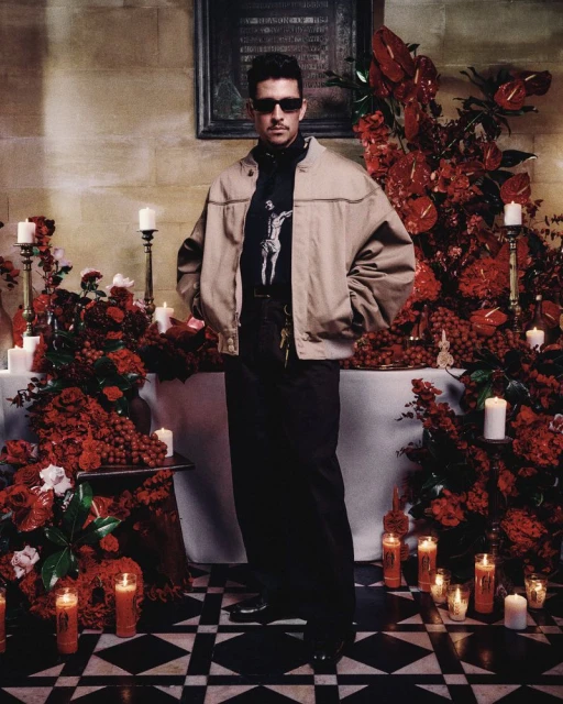 Full shot of a man in a jacket and sunglasses, posing in front of red floral arrangements and candles in a room with tile floors.