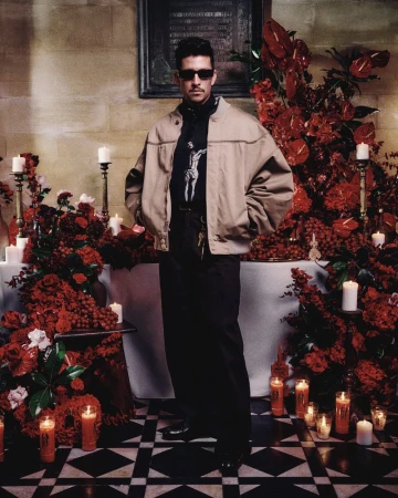 Full shot of a man in a jacket and sunglasses, posing in front of red floral arrangements and candles in a room with tile floors.