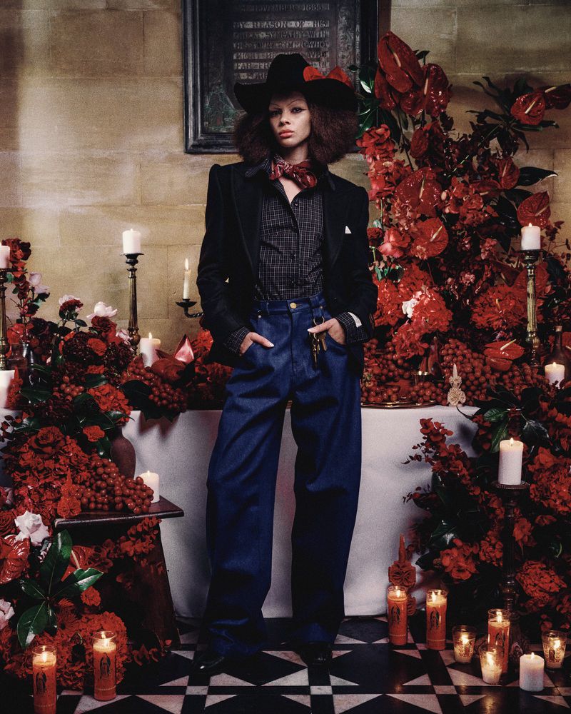 A person in a black blazer, large hat, and blue trousers stands among a red floral display and candles on a black and white floor.