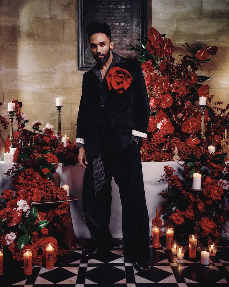 Stylish adult male in a black velvet suit with a large red rose, posing indoors amid red flowers and candles on a black-and-white floor.