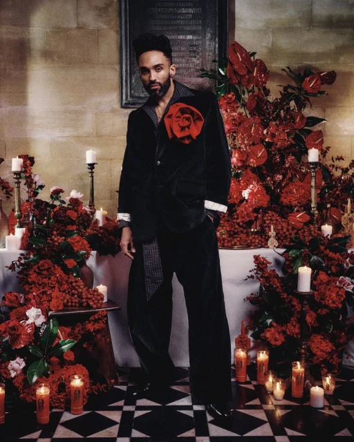 Stylish adult male in a black velvet suit with a large red rose, posing indoors amid red flowers and candles on a black-and-white floor.