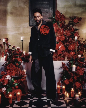 Stylish adult male in a black velvet suit with a large red rose, posing indoors amid red flowers and candles on a black-and-white floor.