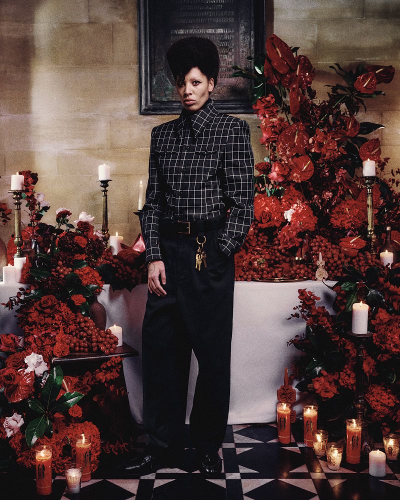 Fashionable individual posing in a church-like setting surrounded by red flowers and candles, wearing a checkered shirt and black pants.