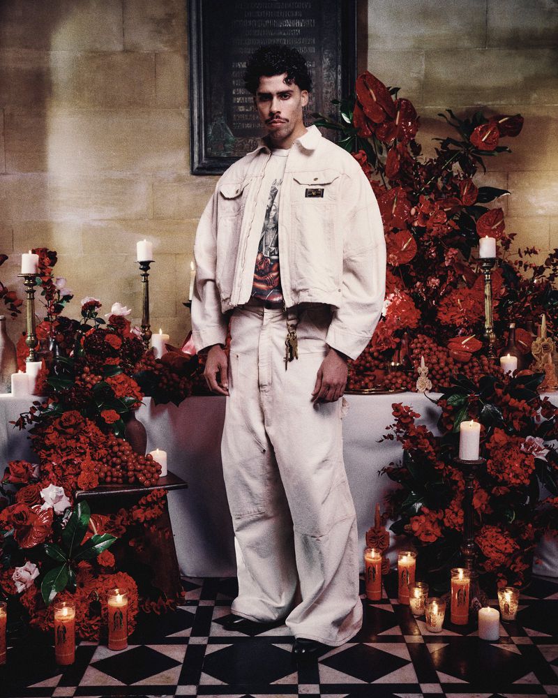Full-length portrait of a young adult male wearing an off-white denim jacket and baggy pants, standing in a room with red flower arrangements.