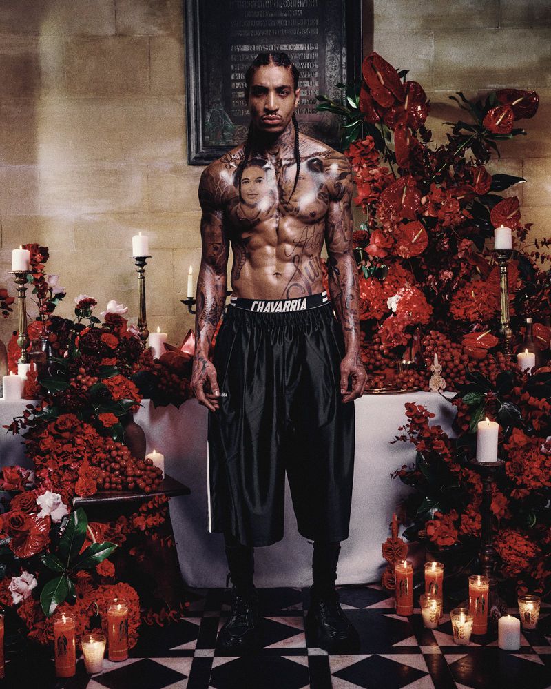 Tattooed man stands in church interior surrounded by candles and red flowers. He is wearing black shorts and a jewelry pendant.