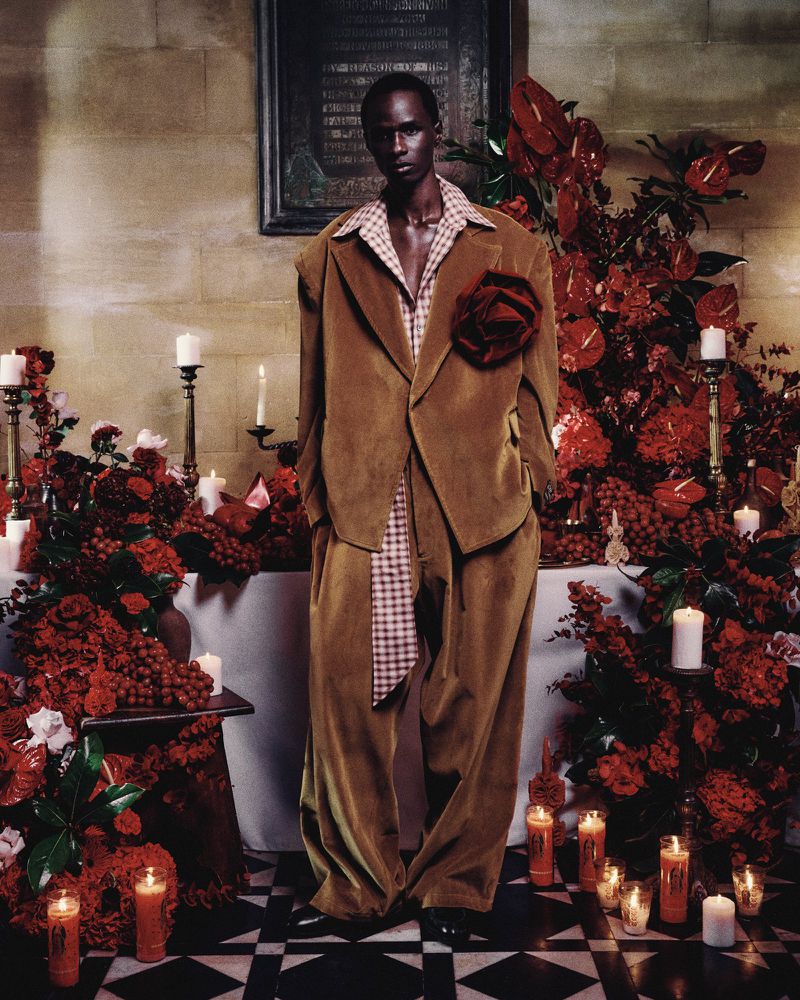 Dark-skinned man in a tan suit with an oversized lapel flower stands in an ornate room with red flowers and lit candles.