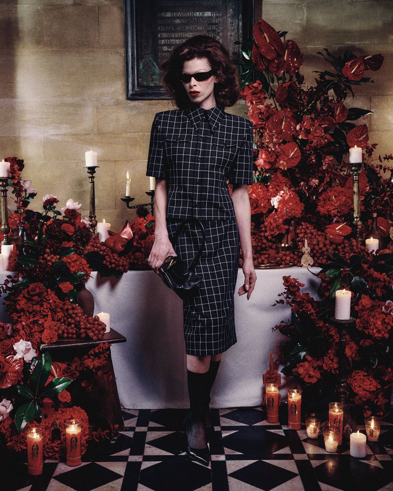 A woman in a checkered outfit poses in an elaborate indoor space with red flowers, candles, and a black-and-white tiled floor.