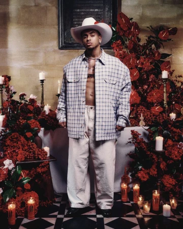 Man in a white cowboy hat and open plaid overshirt posing in front of a red floral candle altar on checkered tile flooring.