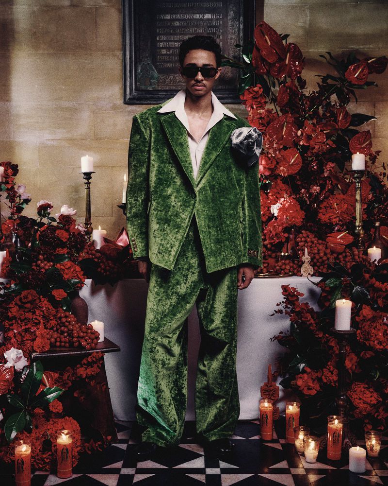 Adult male in a green velvet suit poses indoors surrounded by red flowers and candles, wearing sunglasses and a white collared shirt.