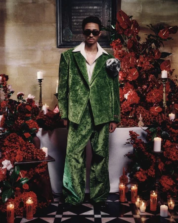 Adult male in a green velvet suit poses indoors surrounded by red flowers and candles, wearing sunglasses and a white collared shirt.