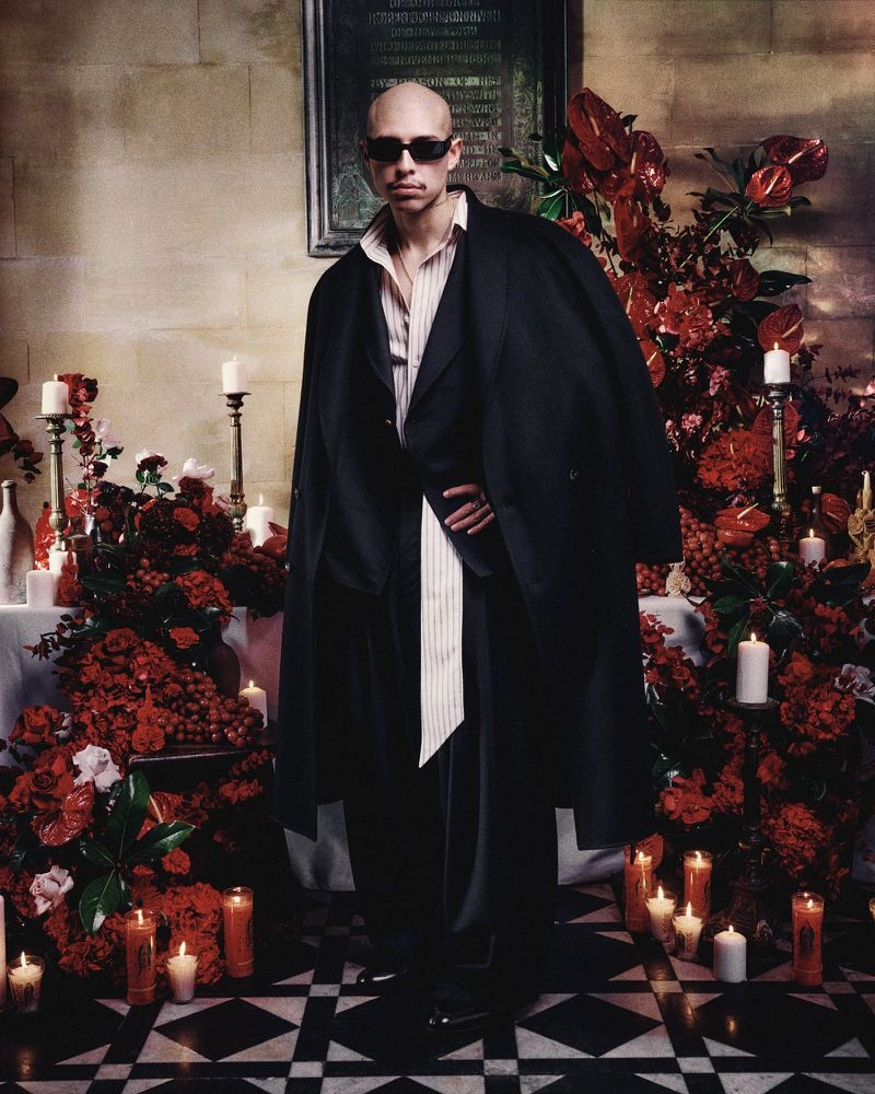 Model wearing dark sunglasses and an oversized black suit with a long draped shirt standing before a candlelit red floral altar.