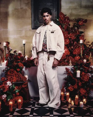 Man wearing cream matching jacket and trousers standing before a table covered in red flowers and lit candles.