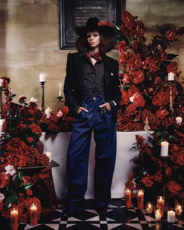 Model wearing a black western hat, black blazer, and wide-leg denim jeans standing amid red floral arrangements and candles.