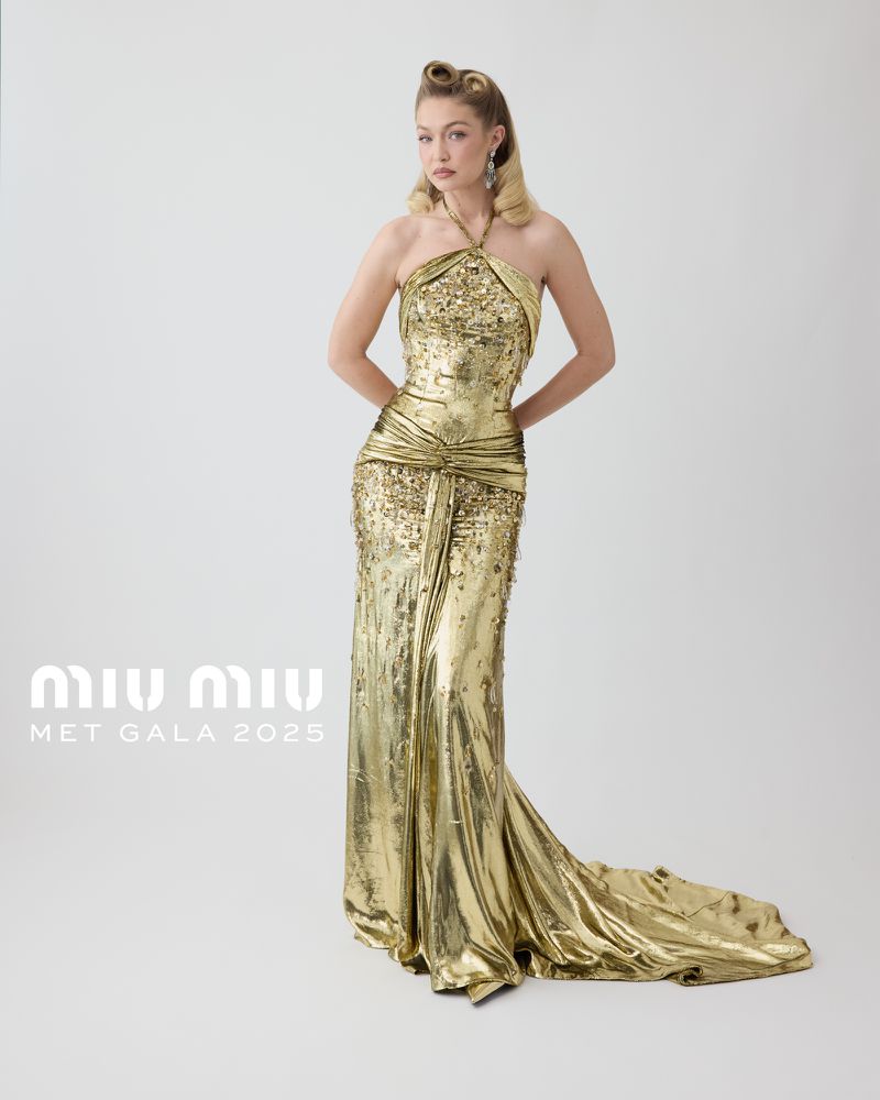 Gigi Hadid in a floor-length gold halter dress. The dress has a train and a fitted bodice. Hadid stands against a white background.