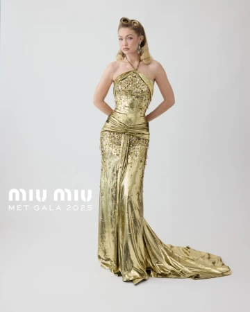 Gigi Hadid in a floor-length gold halter dress. The dress has a train and a fitted bodice. Hadid stands against a white background.