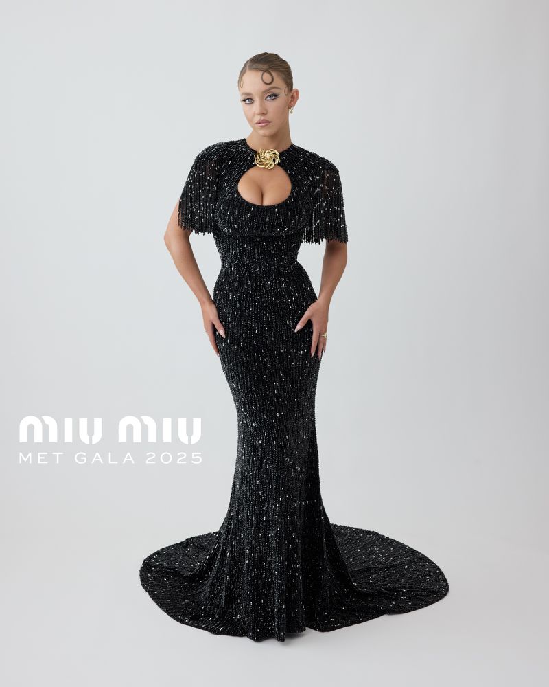 Sydney Sweeney poses in a black, floor-length Miu Miu gown with cape detail and gold brooch against a plain background for the Met Gala 2025.