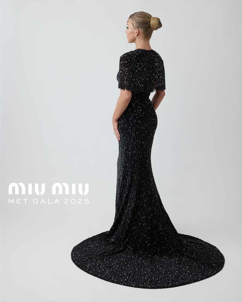Woman in a long black gown and cape, shot from the back, against a white backdrop, promoting Miu Miu's 2025 Met Gala collection.