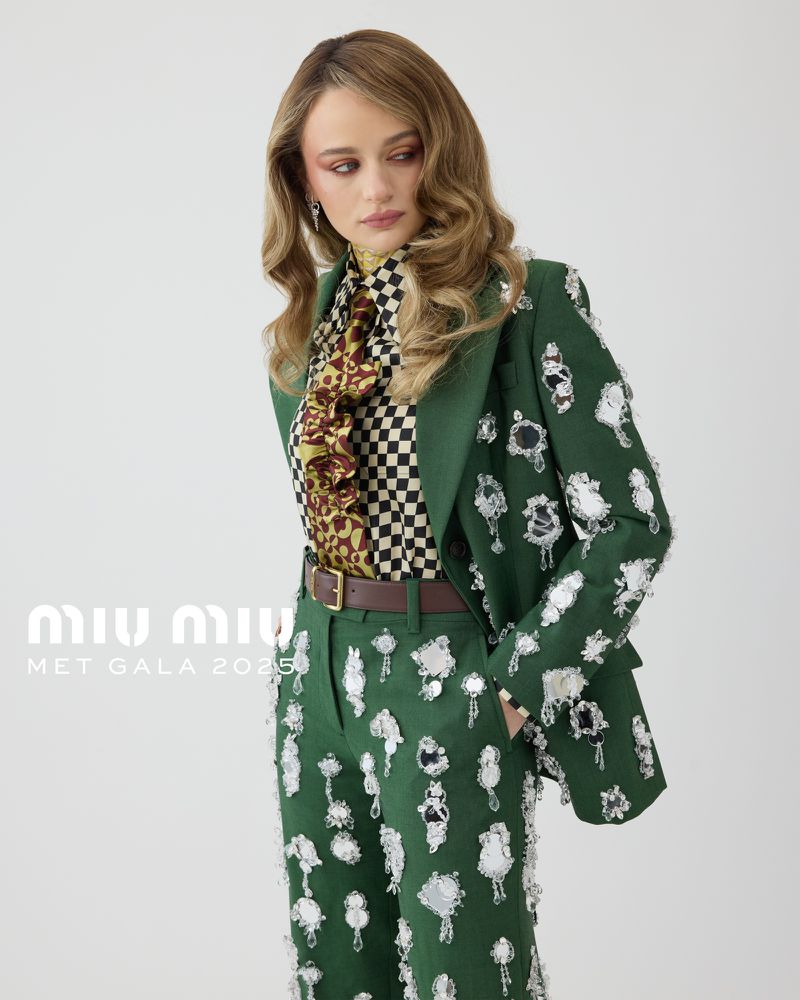 Joey King poses in a green embellished suit with a black and white checkered shirt and brown belt against a white backdrop.