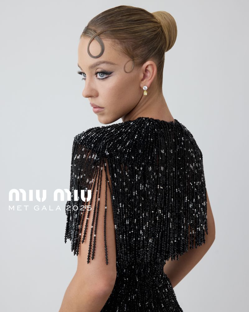 Close-up of a young woman with a high bun and decorative hairstyle wearing a black beaded fringe dress, set against a bright backdrop.