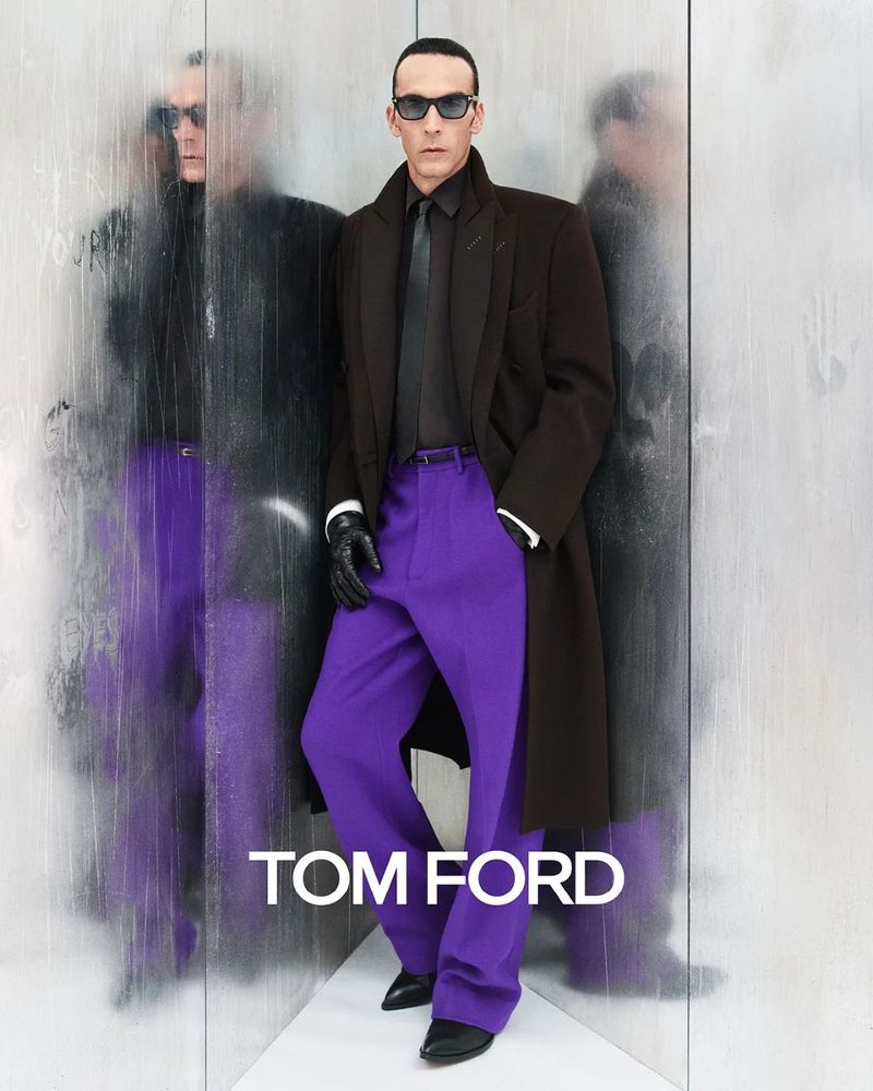 A man in a dark coat and sunglasses poses against a textured silver wall wearing bright purple slacks in a fashion shot.