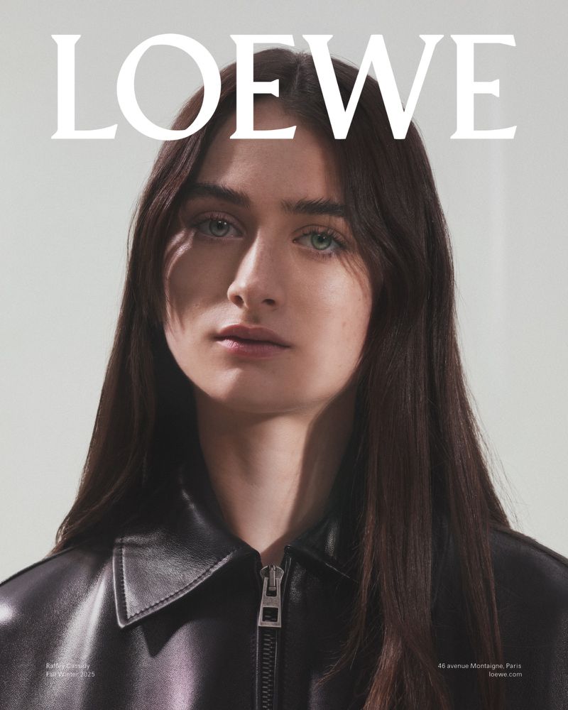 Close-up studio shot of a brunette woman in a black jacket, fashion editorial. White background with Loewe logo and text at bottom.
