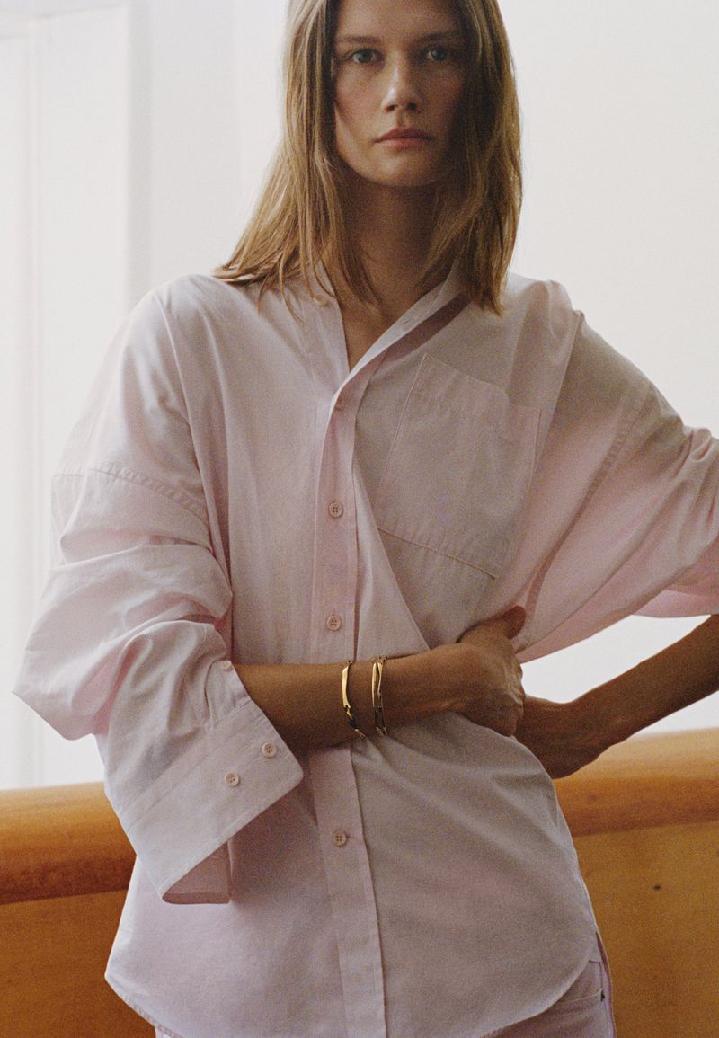 A fair-skinned woman with blonde hair poses indoors wearing a pink button-down shirt and gold bracelets, with soft natural lighting.