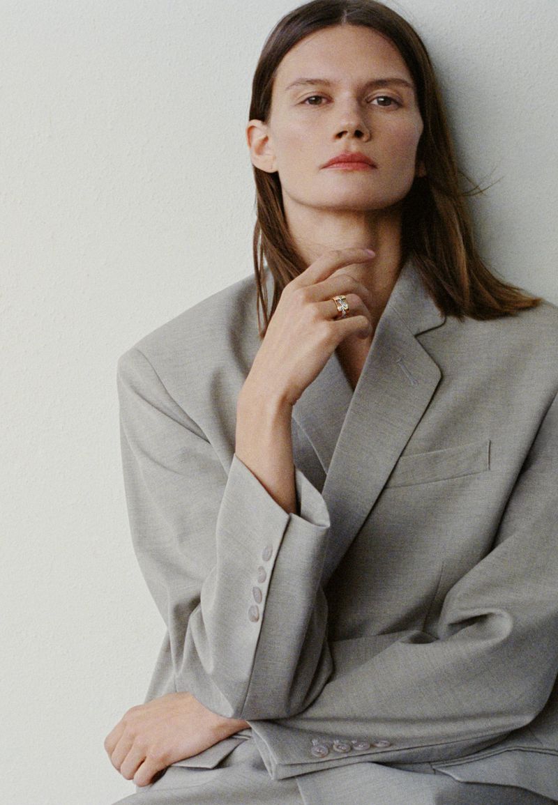 A young woman in a grey blazer poses in a close-up studio shot, wearing a ring on her finger and light makeup against a neutral wall.