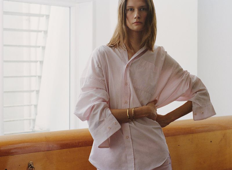 A fashionable woman in a pink button-down shirt poses indoors near a window with sunlight, creating a bright and airy ambiance.