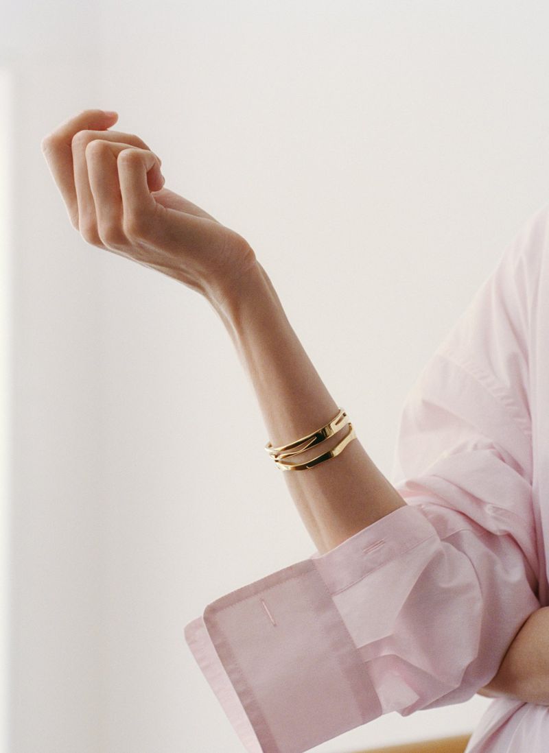 Woman's arm with gold bracelets, wearing a light pink top against a cream background. Jewelry and fashion advertising content.