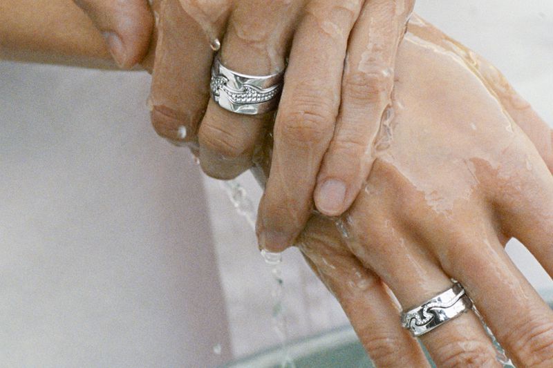 Two hands under running water, showcasing silver rings. Close-up shot of skin, water droplets, and jewelry, hygiene concept.