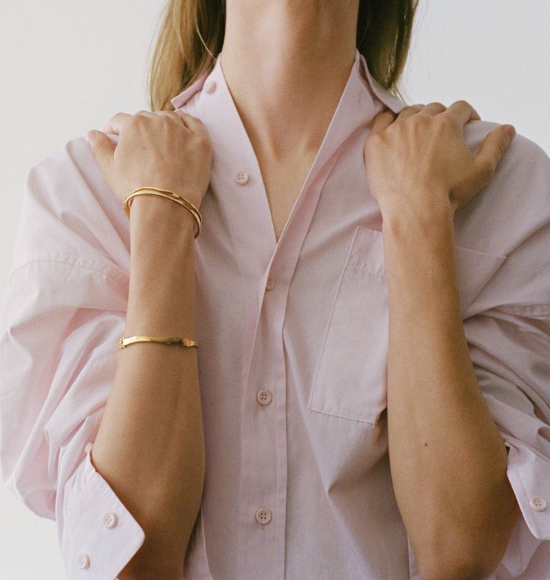 Close-up of a person wearing two gold bracelets with a pink button-down shirt against a white background in a fashion setting.