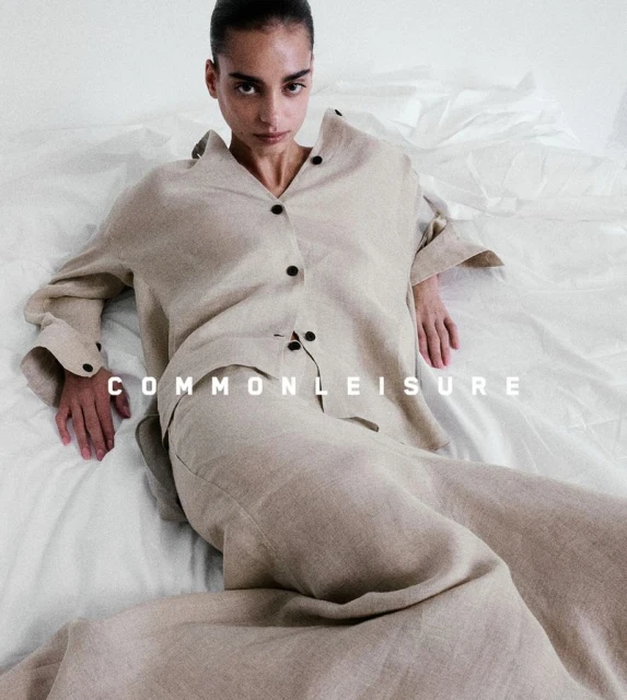 Fashionable adult female model in neutral-toned linen loungewear set posing indoors on white fabric. The image is a fashion editorial.