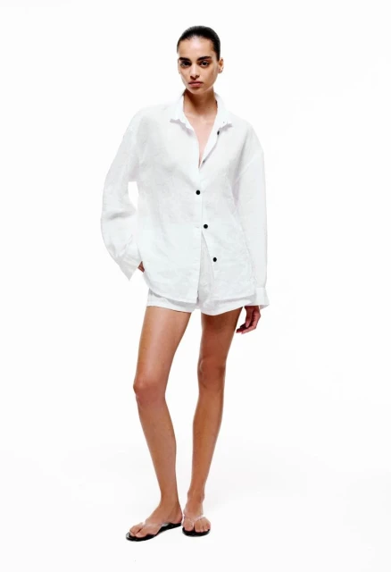 Woman in white oversized shirt and shorts poses in a fashion shot against a white backdrop with a neutral facial expression.