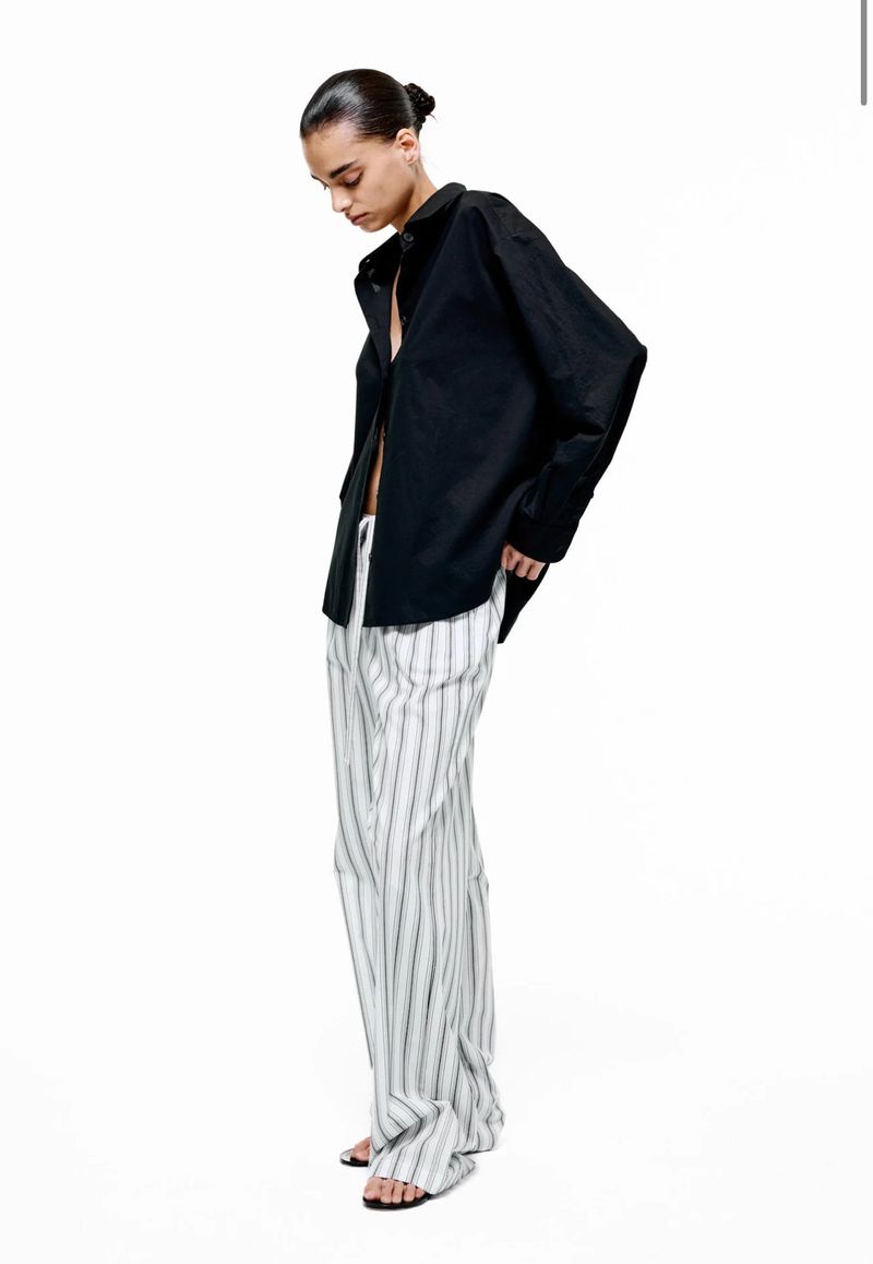 Fashion photo: An adult female model wears a black button-down shirt and striped pants against a white backdrop in a studio shot.