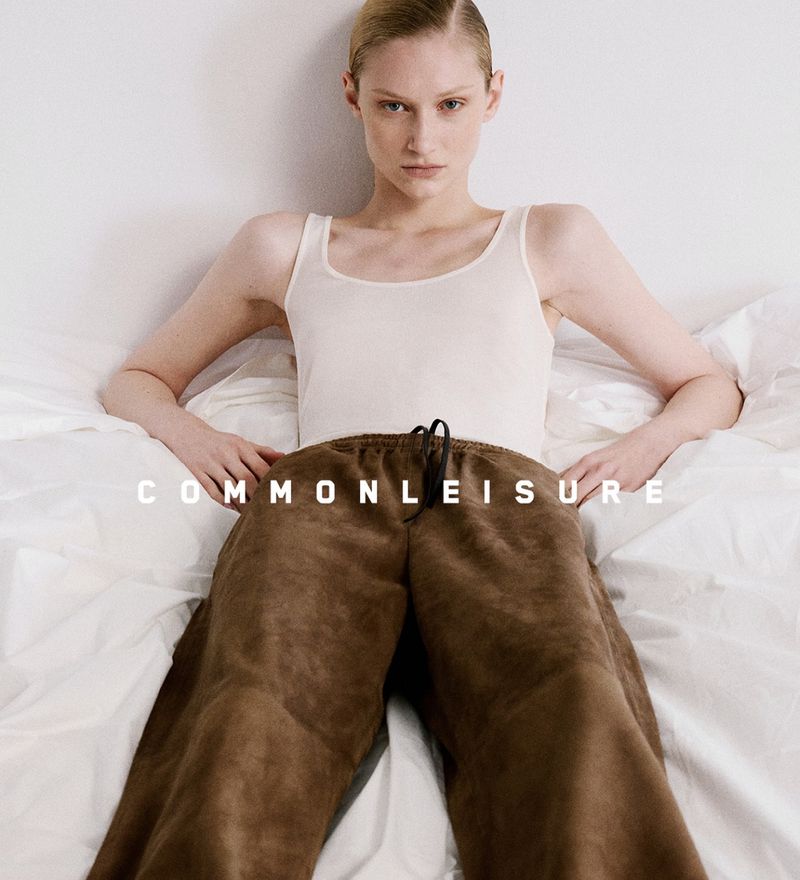 A young fair-skinned adult female model poses in bed wearing a white tank top and brown sweatpants, looking straight at the camera.