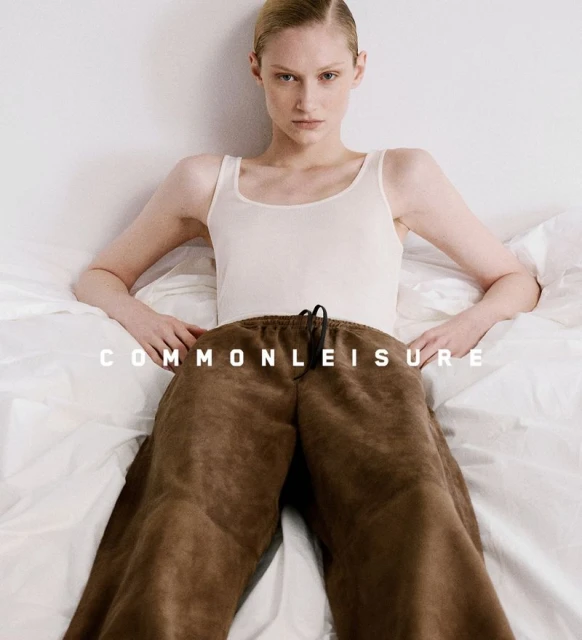 A young fair-skinned adult female model poses in bed wearing a white tank top and brown sweatpants, looking straight at the camera.