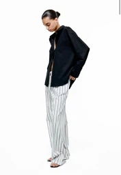 Model posing in an oversized black button-up shirt and wide-leg striped trousers against a white studio background.