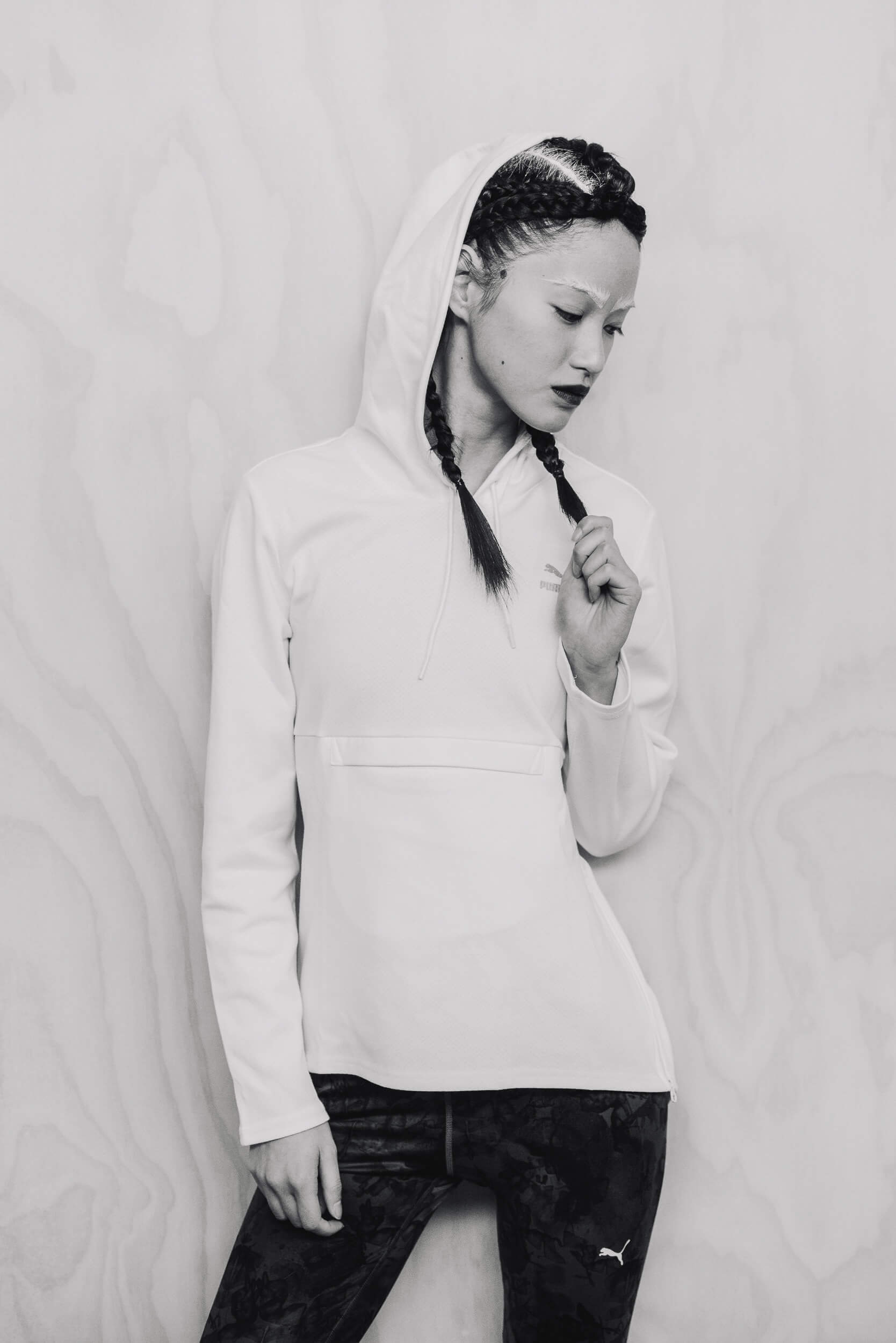 A black and white portrait of a young woman with braided hair, wearing a hooded sweatshirt, posing against a light woodgrain background.
