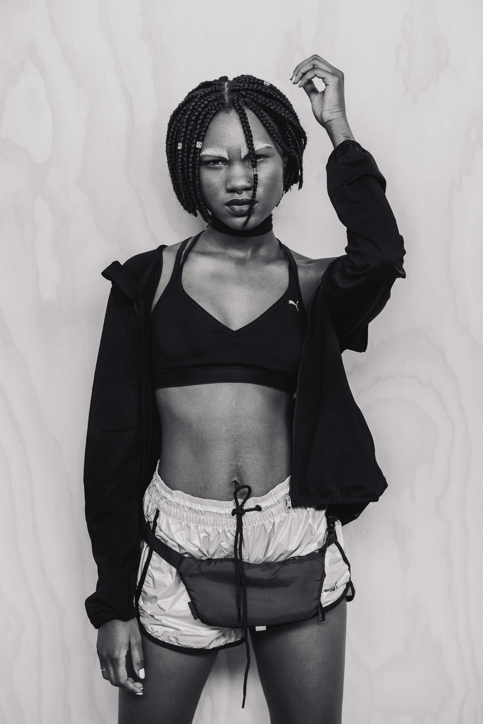 Monochromatic studio shot of a slender Black woman with box braids, wearing a sports bra, shorts, and a fanny pack against a wood wall.