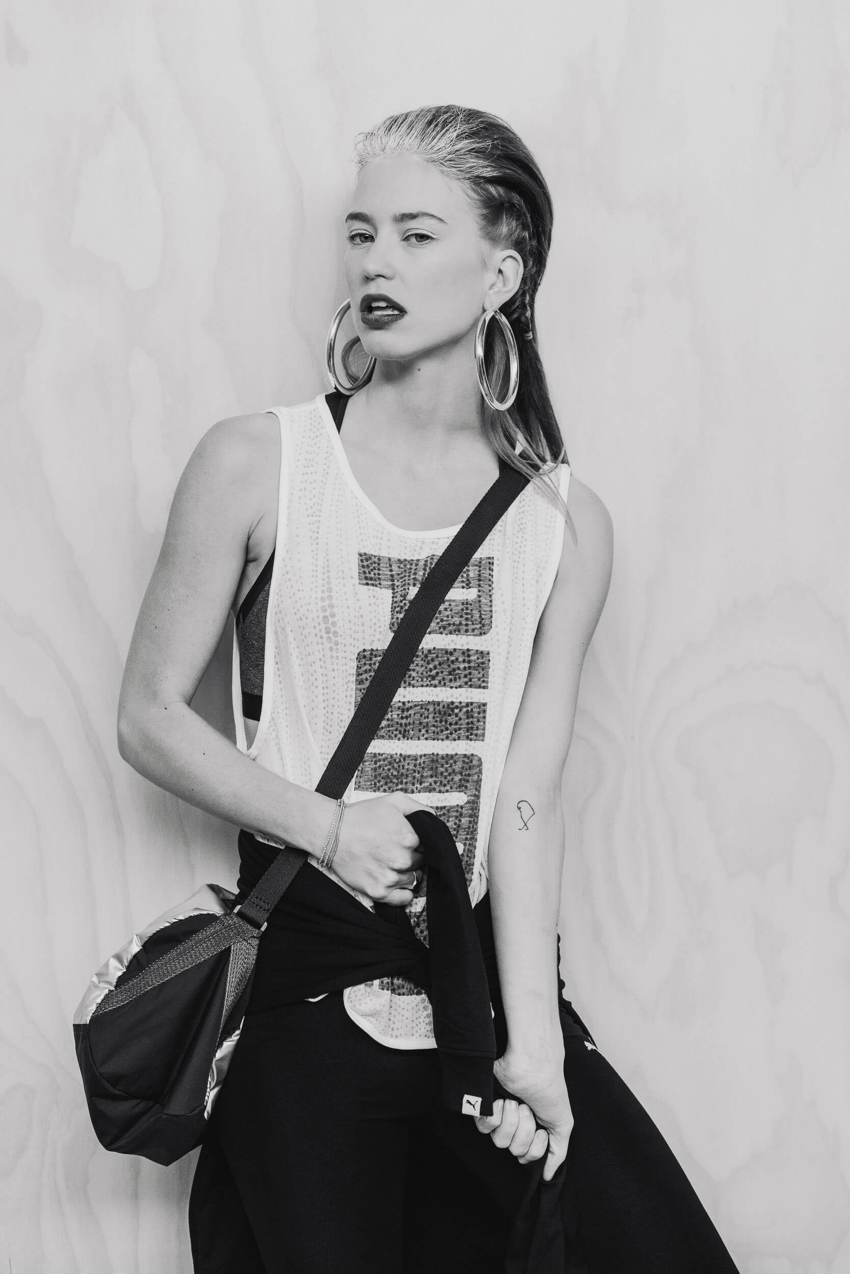 Monochrome portrait of a female model with hoop earrings, wearing a tank top and black pants, and holding a gym bag in a photo studio.