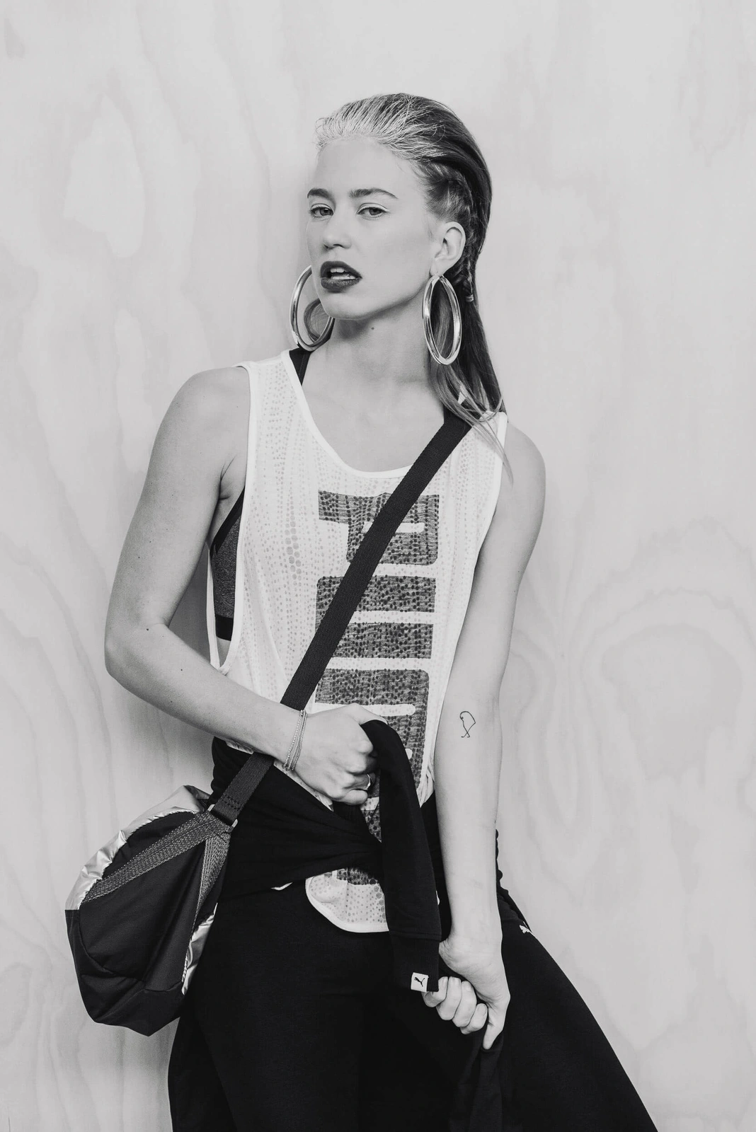 Model in white athletic tank top with large hoop earrings and shoulder bag against a light wood panel background.