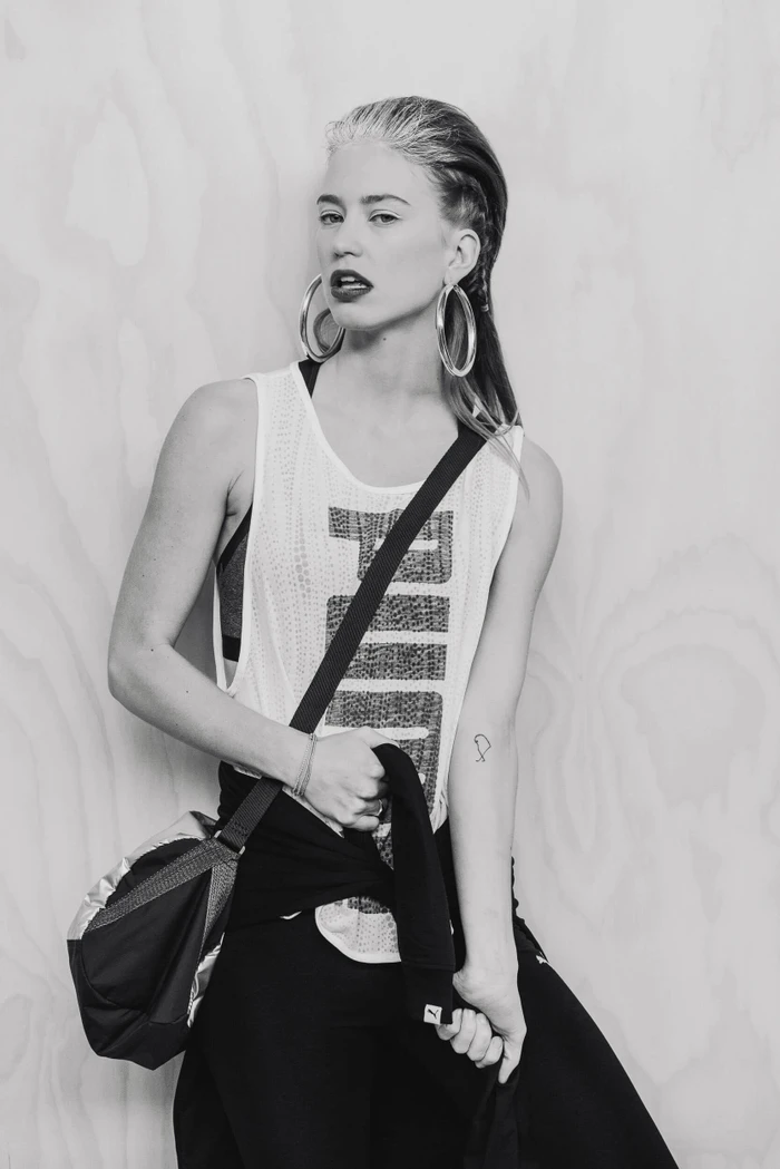 Model in white athletic tank top with large hoop earrings and shoulder bag against a light wood panel background.