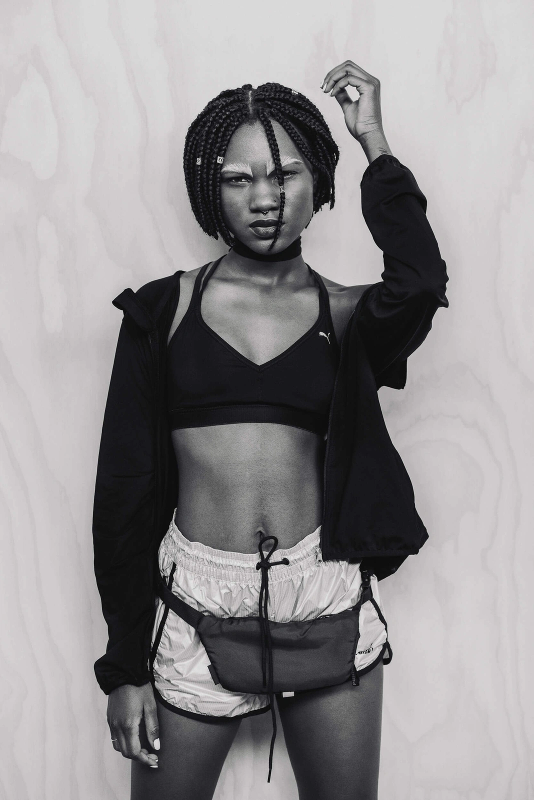 Black and white portrait of a woman wearing Puma sportswear and shorts with a fanny pack against a wood grain wall.