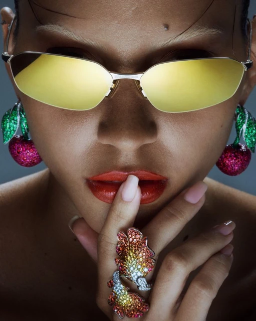 Close-up portrait of a woman with yellow sunglasses, cherry earrings, and statement rings, highlighting fashion and beauty details.